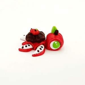 Può includere: Una tartaruga rossa e bianca stampata in 3D con una mela marrone e rossa sulla schiena. La mela ha una foglia verde e un occhio verde.