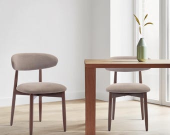 May include: Two dining chairs with dark brown wooden frames and light beige upholstered seats and backrests, positioned next to a rectangular wooden dining table that holds a single green vase with dried decorative grass stems.