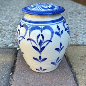 May include: A small, white ceramic jar with a lid, adorned with blue floral patterns. The jar has a rounded shape with a slightly flared base. The lid is also white with a blue border and a decorative blue design. The jar is sitting on a brick surface.