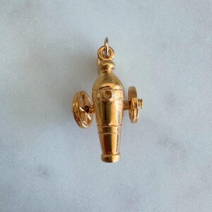 May include: A gold-coloured charm in the shape of a cannon with wheels. The cannon is detailed with a barrel, a base, and two wheels. The charm is designed to be a pendant or a charm for a bracelet.