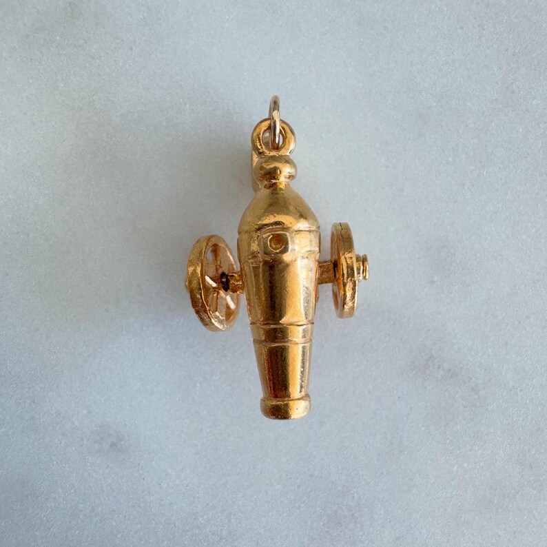 May include: A gold-coloured charm in the shape of a cannon with wheels. The cannon is detailed with a barrel, a base, and two wheels. The charm is designed to be a pendant or a charm for a bracelet.