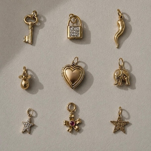 Puede incluir: Una colección de colgantes dorados, que incluyen una llave, un candado con un patrón similar a diamantes, un cuerno, un chupete, un relicario en forma de corazón, un elefante, una estrella con detalles de diamantes, un lazo con una gema roja y una estrella texturizada.