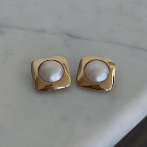 Könnte beinhalten: Ein Paar goldfarbene quadratische Ohrringe, jeweils mit einem großen, schillernden, weißen Perlenmittelstück. Die Ohrringe haben ein klassisches, elegantes Design, das für formelle oder alltägliche Anlässe geeignet ist.