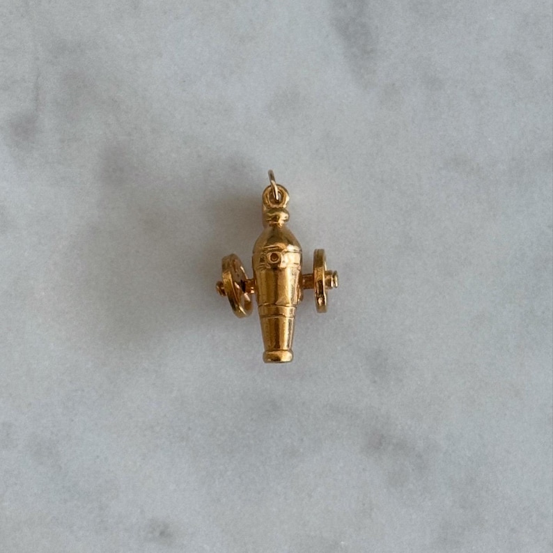 May include: A gold-coloured charm in the shape of a cannon with wheels. The cannon is detailed with a textured surface and a small opening at the end.