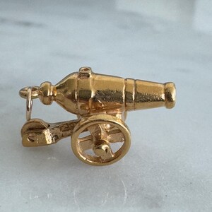 May include: A gold-coloured charm in the shape of a cannon. The cannon has a wheel on one side and a barrel that points to the right. The charm is detailed and has a realistic design.