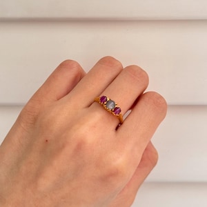 Pode incluir: Um anel de ouro com três pedras. A pedra central é um oval cinza claro, e as duas pedras de cada lado são ovais rosa escuro.