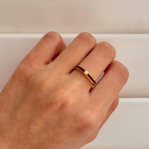 Pode incluir: Um anel de ouro com uma faixa de esmalte vermelho escuro e uma pequena pedra preciosa transparente. O anel é mostrado no dedo de uma pessoa, destacando o design e o artesanato. O anel é uma joia.