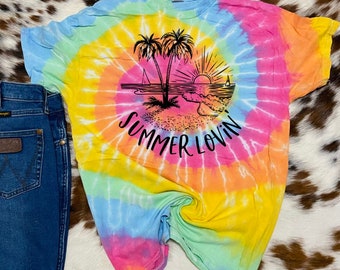 Puede incluir: Una camiseta tie-dye con un esquema de color arcoíris. La camiseta presenta un gráfico en blanco y negro de palmeras, una puesta de sol y el texto "Summer Lovin'"