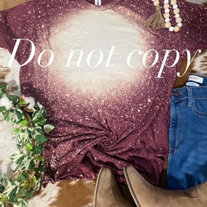May include: A maroon t-shirt with a white bleach design. The shirt is made of a soft, comfortable material and has a relaxed fit. The shirt is perfect for everyday wear.