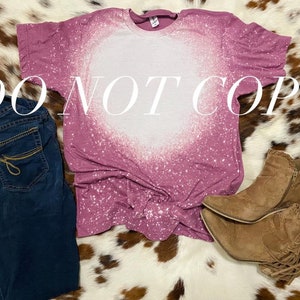 May include: A mauve t-shirt with a white bleach design in the shape of a heart. The shirt is on a white and brown cowhide background with a pair of brown suede fringe boots.