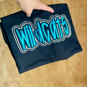 May include: A folded black t-shirt with the word "Wildcats" in a teal, outlined, bubble letter design. The text is bordered by a silver dotted line. The shirt is held up by a hand.