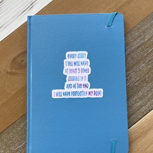 Puede incluir: Un cuaderno azul con una pegatina que dice "Cada historia que cuento tendr&aacute; al menos 5 historias m&aacute;s en ella y al final me habr&eacute; olvidado de mi punto."