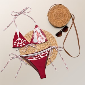 Könnte beinhalten: Rotes Bikini-Set mit weißen Akzenten und einem einzigartigen Design. Das Oberteil hat eine Dreiecksform mit Muster, die Hose ein passendes Design. Eine gewebte Tasche, Sonnenbrille und eine Strohmatte sind ebenfalls im Bild.