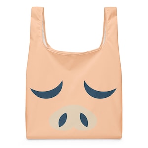 Puede incluir: Una bolsa de compras reutilizable de color melocotón con un diseño de cara de cerdo. La cara del cerdo incluye líneas curvas azul oscuro para los ojos y un hocico beige claro con orificios nasales azul oscuro. La bolsa tiene un interior blanco.