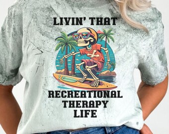 May include: A light gray t-shirt with a faded design of a skeleton wearing sunglasses and riding a surfboard. The text "Livin' That Recreational Therapy Life" is printed on the shirt.