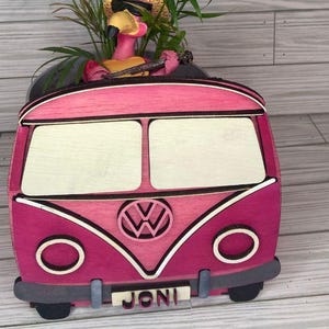 May include: Pink wooden Volkswagen bus planter with a flamingo figurine sitting inside. The bus has a white VW logo and the name "Joni" on the front.