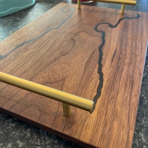 May include: A rectangular wooden serving tray with brass handles. The tray is made of dark brown wood with a black resin river design. The brass handles are cylindrical and mounted on the short sides.