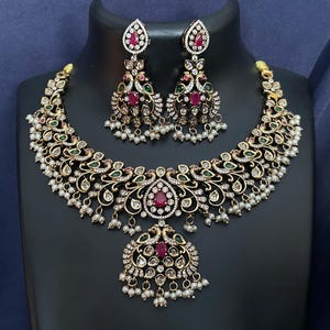 May include: A gold-toned jewelry set featuring a necklace and matching earrings. The necklace has a central pendant with a red gemstone and is adorned with white, green, and red stones. The earrings mirror the necklace design, with dangling white pearls.