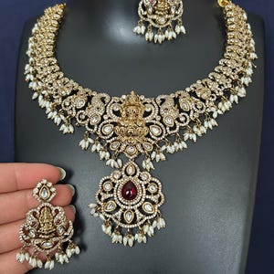 May include: A gold-toned necklace and earring set. The necklace features a central pendant with a large, teardrop-shaped red stone, surrounded by clear stones and small white pearls. The earrings mirror the necklace design. The set is displayed on a black surface.