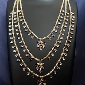 May include: A multi-strand necklace with a pearl-like bead design. Each strand features teardrop-shaped clear crystals and a floral pendant with red gemstones. The necklace is displayed on a black mannequin.