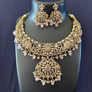 May include: A gold-toned necklace and earring set with intricate filigree and pink accents. The necklace features a central pendant and matching earrings, all adorned with small pink beads and clear crystals. The set is displayed on a black mannequin.