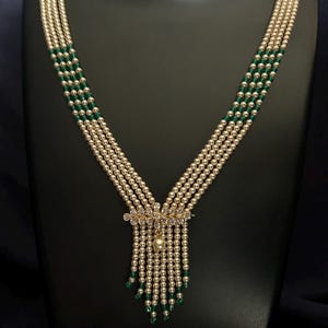 May include: A multi-strand necklace featuring faux pearls and green beads. The necklace has a gold-colored floral pendant with clear accents and a single pearl drop. The necklace is displayed on a black mannequin.