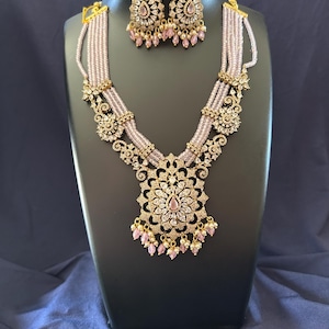 May include: A gold-toned necklace and earring set displayed on a black mannequin. The necklace features multiple strands of light pink beads, ornate gold filigree, and a large pendant with a pink center stone. The earrings match the necklace with similar detailing.