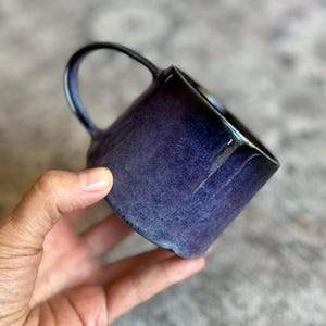 May include: A ceramic mug with a deep indigo and violet glaze featuring a mottled, textured finish. The mug has a rounded body and a single curved handle extending from the side, showcasing a dark, glossy rim and a slightly irregular, organic shape.