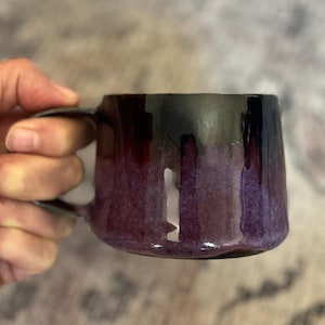 May include: A ceramic mug featuring a glossy glaze that transitions from a deep, dark black at the rim to a vibrant, mottled purple at the base. The mug has a wide, rounded body and a sturdy, curved handle attached to the side of the vessel.