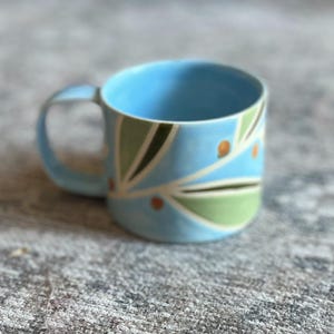 May include: A light blue ceramic mug featuring a painted pattern of green leaves and small orange dots. The mug has a wide, cylindrical body and a curved handle on the side, with a smooth finish and a consistent color palette throughout the design.