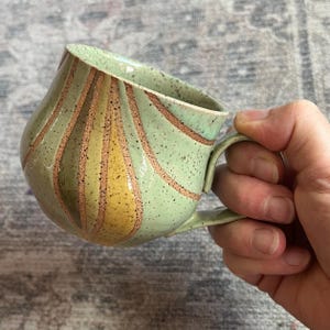 May include: A ceramic mug with a speckled light green glaze featuring vertical, carved-out panels in shades of yellow and brown. The mug has a rounded, bulbous body and a curved handle extending from the side, showcasing a textured finish.
