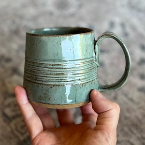 May include: A handmade ceramic mug with a seafoam green glaze. The mug features horizontal textured lines around the middle and a rounded handle. The base of the mug is a natural, unglazed clay color. A unique piece for enjoying coffee or tea.