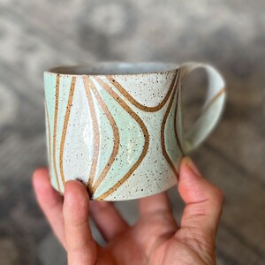 May include: A ceramic mug with a speckled off-white glaze featuring curved, light mint green panels separated by thin, dark brown lines. The mug has a wide, flat-bottomed cylindrical shape and a curved handle extending from the right side.