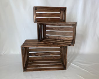 May include: Three rectangular wooden crates with a dark brown stained finish, stacked in an offset arrangement. Each crate features horizontal slats on the sides and bottom, with solid corner supports visible at the edges of the open box structure.