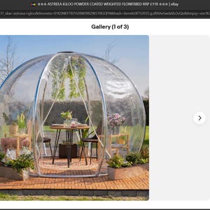 May include: A clear, dome-shaped outdoor structure with a table and chairs inside. The structure is surrounded by plants and flowers, creating a garden-like setting. The frame is silver and the dome is transparent. The structure is on a wooden deck.