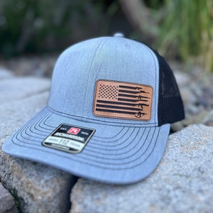 May include: A gray and black trucker hat with a brown leather patch featuring an American flag with guns. The patch has the text "EST. 1970" on it.