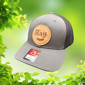 May include: Gray baseball cap with a black mesh back and a curved brim. A circular wooden patch on the front features the word "Stay" above "One More Day" and a feather graphic. The cap has a Flexfit tag.