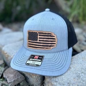 May include: A gray and black trucker hat with a brown leather American flag patch on the front. The hat has a black mesh back and a black adjustable strap with a label that reads "Est. 1970 Richardson 112 Original Trucker Adjustable".