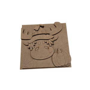 May include: A light brown, flat, laser-cut wooden craft cutout featuring a moose wearing a hat with a plaid pattern and a small tag, positioned next to a pumpkin. The design is layered on a square base, showcasing a smooth, unfinished wood texture.