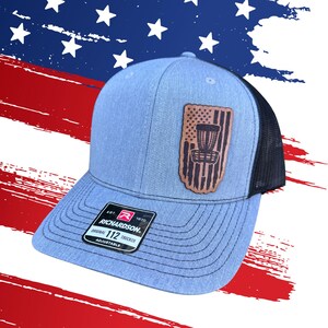 May include: A blue trucker hat with a black mesh back and a brown leather patch featuring a disc golf basket design within an American flag. The hat has a curved brim and the brand name "RICHARDSON" is visible.