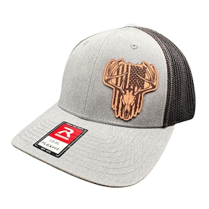 May include: Gray baseball cap with a brown leather patch featuring a deer skull and American flag design. The cap has a black mesh back and a curved brim. The Richardson brand logo is visible on the front.