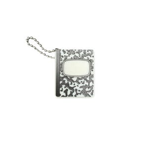 May include: A rectangular metal keychain featuring a white background with a raised, dark gray, irregular camouflage-like pattern. The item includes a vertical textured spine on the left side and a central white oval plaque, attached to a silver ball chain.
