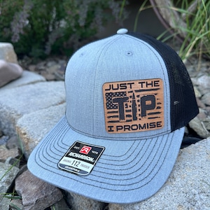 May include: A gray and black trucker hat with a brown leather patch that says "Just the Tip I Promise" with an American flag and a bullet graphic. The hat is adjustable and has a Richardson 112 label.