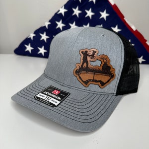 May include: A gray and black trucker hat with a brown leather patch featuring a silhouette of a person with a bow and arrow. The patch is shaped like a state of Texas. The hat has a Richardson 112 Trucker label.