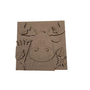 May include: A square light brown wooden cutout featuring a layered moose design. The moose is depicted with large antlers, a rake held in one hoof, and several detailed leaf shapes scattered around the head and body. The surface is flat and matte.