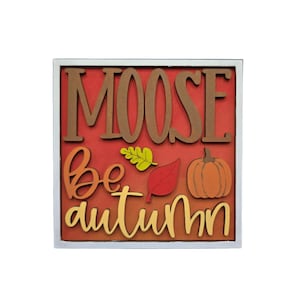 May include: A square wooden sign with a white frame featuring the text MOOSE be autumn in layered brown, orange, and yellow lettering. The design includes a small yellow leaf, a red leaf, and an orange pumpkin on a solid red background.
