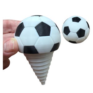 May include: A white and black soccer ball-shaped bottle stopper with a spiral base. The stopper is held by a hand. A second, smaller soccer ball is also visible.