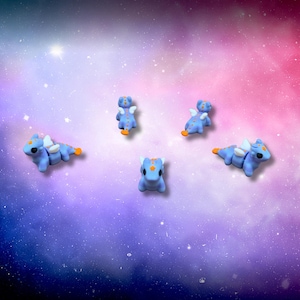 May include: Five small, light blue dragon figurines with white wings and orange accents are arranged against a colorful, galaxy-themed background. The dragons are in various poses, some standing and others lying down.