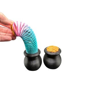 May include: A colorful rainbow slinky toy arches over a black pot, with another pot filled with gold coins. The slinky transitions from pink to blue. The pots are black and round, evoking a St. Patrick's Day theme.