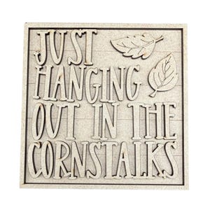 May include: A square wooden sign with a light tan finish featuring the laser-cut text JUST HANGING OUT IN THE CORNSTALKS in a bold, sans-serif font. Two small, detailed leaf cutouts are positioned in the upper right corner of the sign.